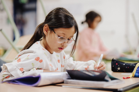 Une jeune fille de 7 ans avec des lunettes réalise un exercice dans son cahier. Elle est dans une salle de classe.
