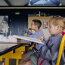 Une petite fille blonde lit un livre dans une classe d'école maternelle. Un autre élève derrière elle est concentré sur le tableau.