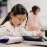Une jeune fille de 7 ans avec des lunettes réalise un exercice dans son cahier. Elle est dans une salle de classe.